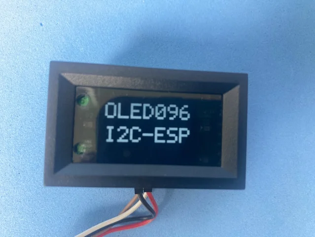 Panel-mount OLED module with wires clipped into screw terminals, screen showing 'OLED096 I2C-ESP', installed in a black enclosure against a light blue background