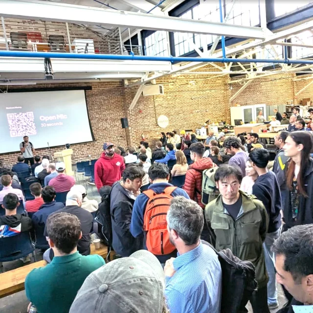 Packed indoor talk at AI for Science Summit — exposed brick walls, steel-frame loft space, screen showing "Open Mic 30 seconds"