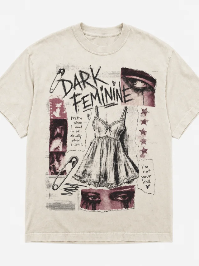 Dark Feminine Babydoll tee — zine-style collage with safety pins, babydoll dress line art, handscrawled "Dark Feminine" text, faded rose and charcoal on cream