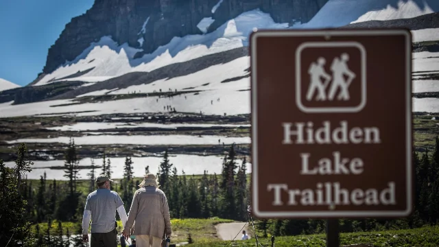 Two hikers walking toward a brown "Hidden Lake Trailhead" sign; snow-patched mountain peaks rise behind them under blue sky