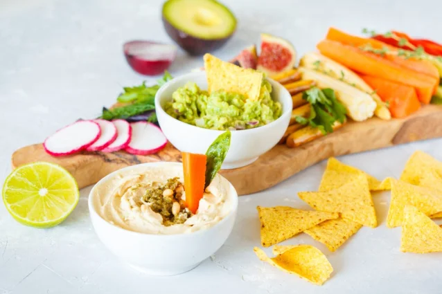 A board of hummus, avocado, vegetables, fruit, and crunchy snacks