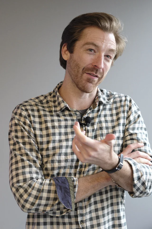 Rob Fitzpatrick, author of The Mom Test, speaking with hands gesturing, wearing a plaid flannel shirt, lavalier microphone, grey background