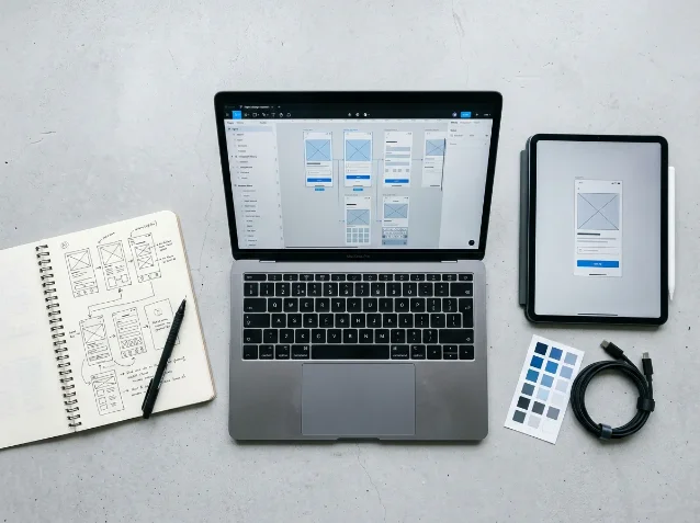A UI/UX designer's workspace with a laptop showing a Figma wireframe canvas and component panel, a spiral notebook with hand-drawn wireframe sketches, a color palette swatch card, and a tablet displaying a mobile UI mockup