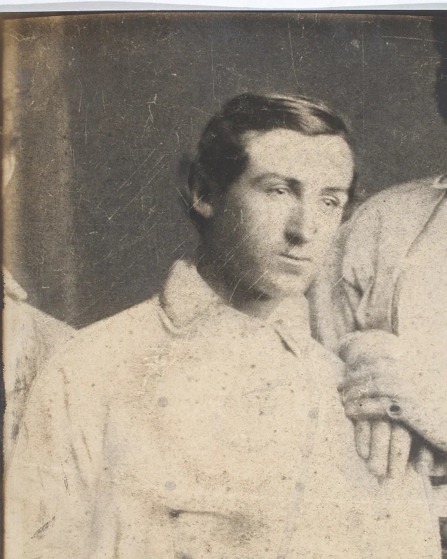 A sepia photograph of a young clean-shaven man in a white collared shirt, looking off to one side, with a teammate's arm and shoulder at the right edge of the frame
