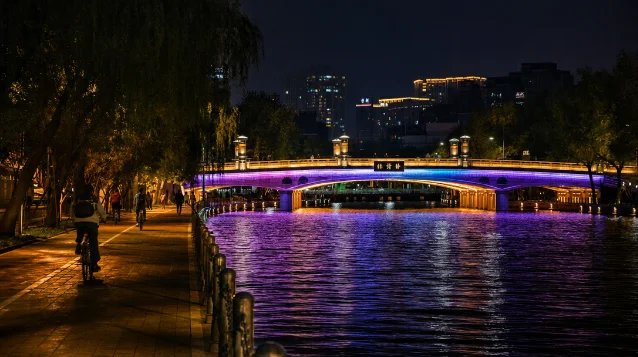 亮马河夜间桥梁灯光,色彩斑斓的 LED 灯带倒映水面,岸边骑行者剪影