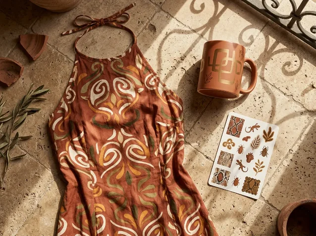 Tuscan Mom Aesthetic product concept — terracotta-scroll printed halter dress, gold-motif mug, and medallion sticker sheet on travertine tile