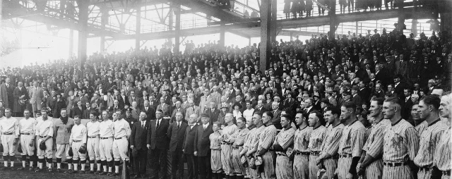 A wide black-and-white photograph of two baseball teams standing in long lines on a dirt field in front of a packed double-decked grandstand, with a small group of men in dark suits and hats at the centre.