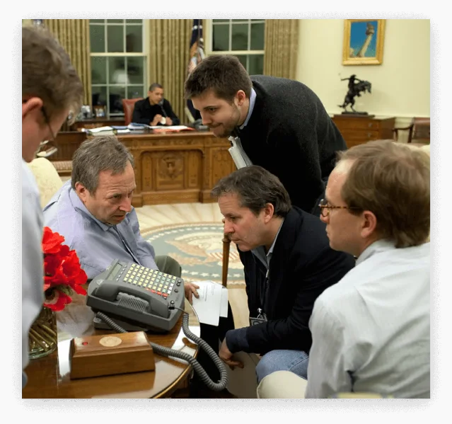 Auto Task Force advisors huddle over documents at the Oval Office while President Obama works at his desk in the background