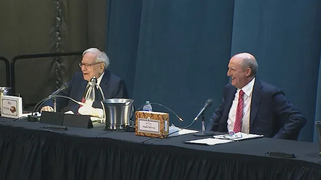 Warren Buffett and Greg Abel seated at a conference table, peanut brittle box visible, both smiling