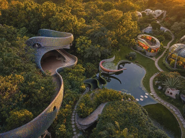 Aerial view of an organic-architecture park with serpentine mosaic buildings, ponds, and stained glass structures at golden hour