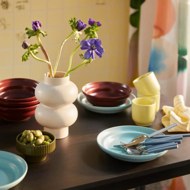 SPJUTLILJA tableware scene — red and dark-red stacked bowls, yellow mugs, sky-blue plates with cutlery on a dark wood table, styled with a white vase and dish towel