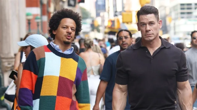 Eric André (left, in a colorful patchwork sweater) and John Cena (right, in a black polo) walking through a crowded New York City street in a scene from Little Brother