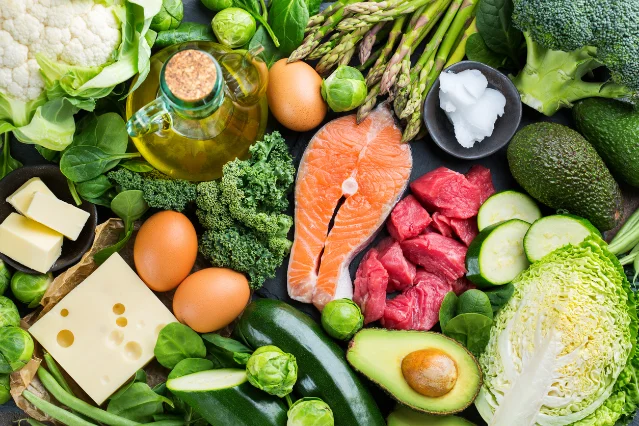 An overhead flat-lay of keto diet ingredients: salmon, raw beef, eggs, avocado, leafy greens, olive oil, butter, cheese, and coconut oil