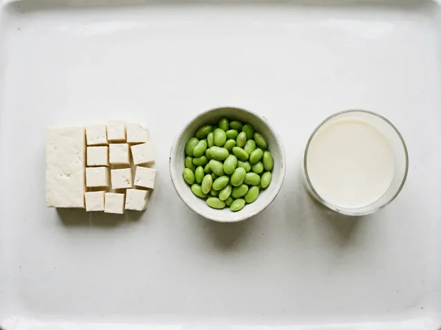 Flat-lay of firm tofu cubes, a bowl of shelled edamame, and a glass of soy milk on a white surface — the three most practical dietary sources of isoflavones in the optimal intake range identified by the study