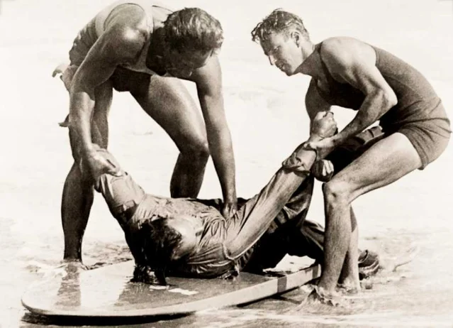 Duke Kahanamoku and Bill Herwig demonstrate the surfboard rescue technique, with Owen Hale playing the role of the rescued victim, in shallow water shortly after the event