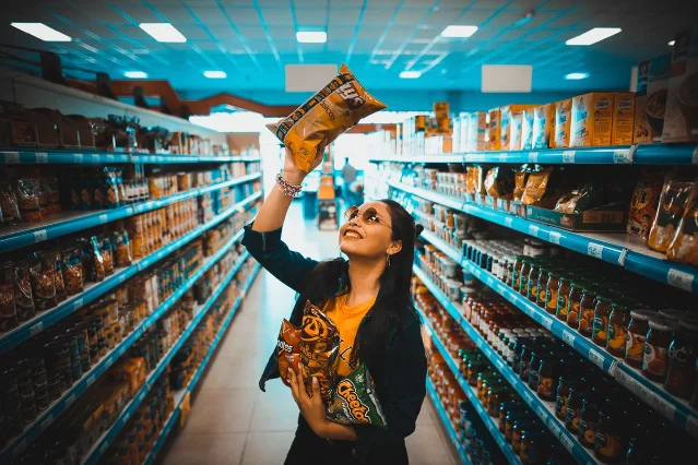A woman in a supermarket holds up two bags of chips, smiling