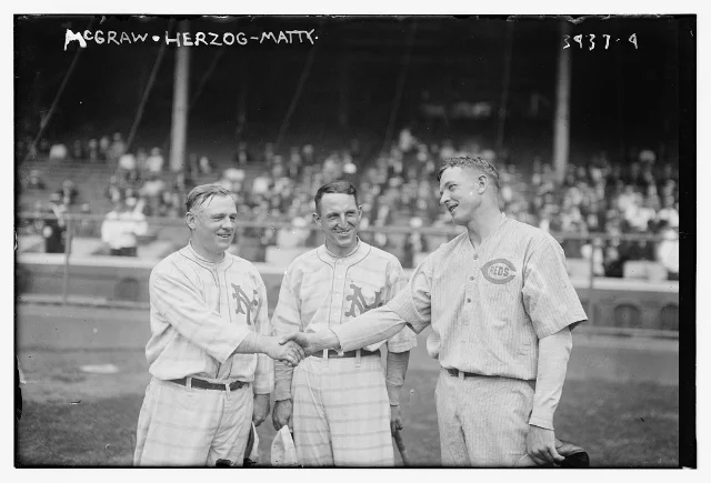 Three men in baseball uniforms shake hands in front of a crowded grandstand in 1916: John McGraw of the Giants on the left, Buck Herzog in the middle and Christy Mathewson in a Cincinnati uniform on the right