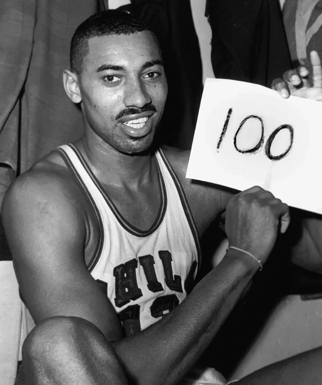 A man in a basketball jersey with dark trim seated and holding up a sheet of paper with 100 written on it