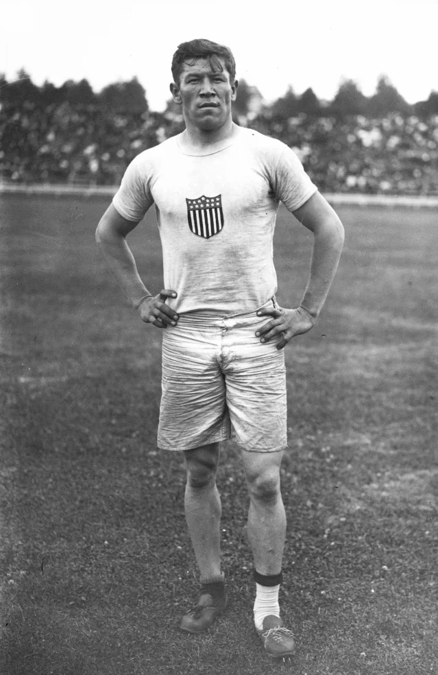A black-and-white full-length photograph of a muscular athlete standing on grass in a white sleeveless jersey with a shield emblem, hands on his hips, wearing two different running shoes