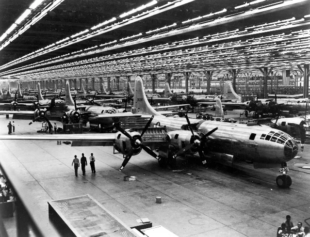 Interior of Boeing Wichita Plant II in 1944, showing B-29 Superfortress bombers in various stages of assembly on the production line under brilliant fluorescent lights, with workers dwarfed by the aircraft