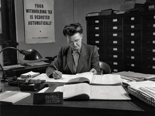 A payroll section clerk at her desk, 1940s, with a "Your Withholding Tax Is Deducted Automatically" government poster on the wall behind her