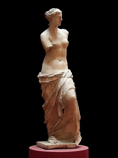 Three-quarter view of the Venus de Milo against a black background, showing the full interplay of the polished torso and deeply carved drapery