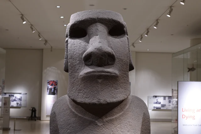 Front view of Hoa Hakananai'a showing the heavy brow, prominent nose, hollowed eye sockets, and the "Living and Dying" gallery visible in the background at the British Museum