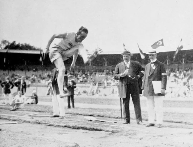 A black-and-white photograph of an athlete in white kit airborne over a sand pit with a crowd and officials watching