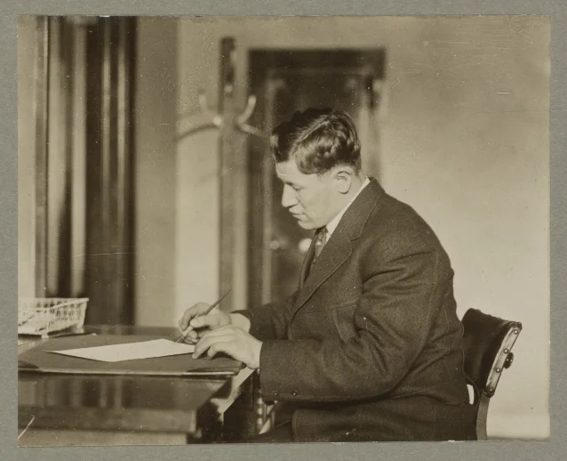 A sepia photograph of a young man in a jacket and tie seated at a desk, pen in hand over a sheet of paper, in an office