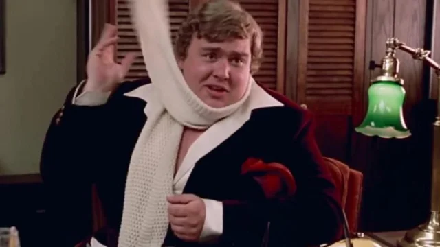 John Candy in a film still from I Like Me — seated indoors, wearing a black coat and loosening a white knit scarf, looking expressive