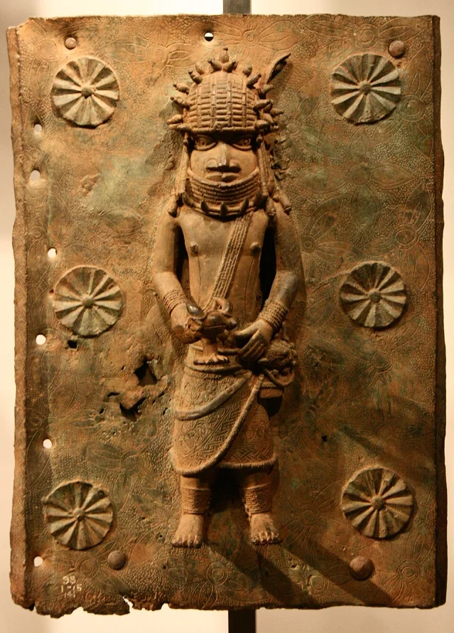 Benin brass plaque showing a standing warrior figure with elaborate crown regalia and river-leaf background pattern